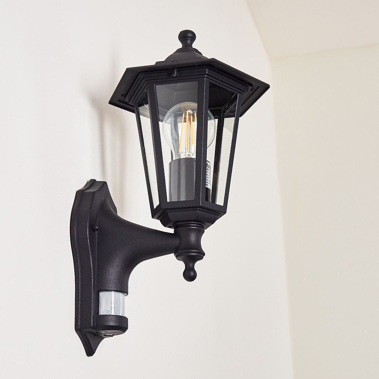 Eine schwarze Außenwandleuchte mit Glasgehäuse und Glühbirne. Sie ist an einer weißen Wand montiert. Hat einen Bewegungssensor.