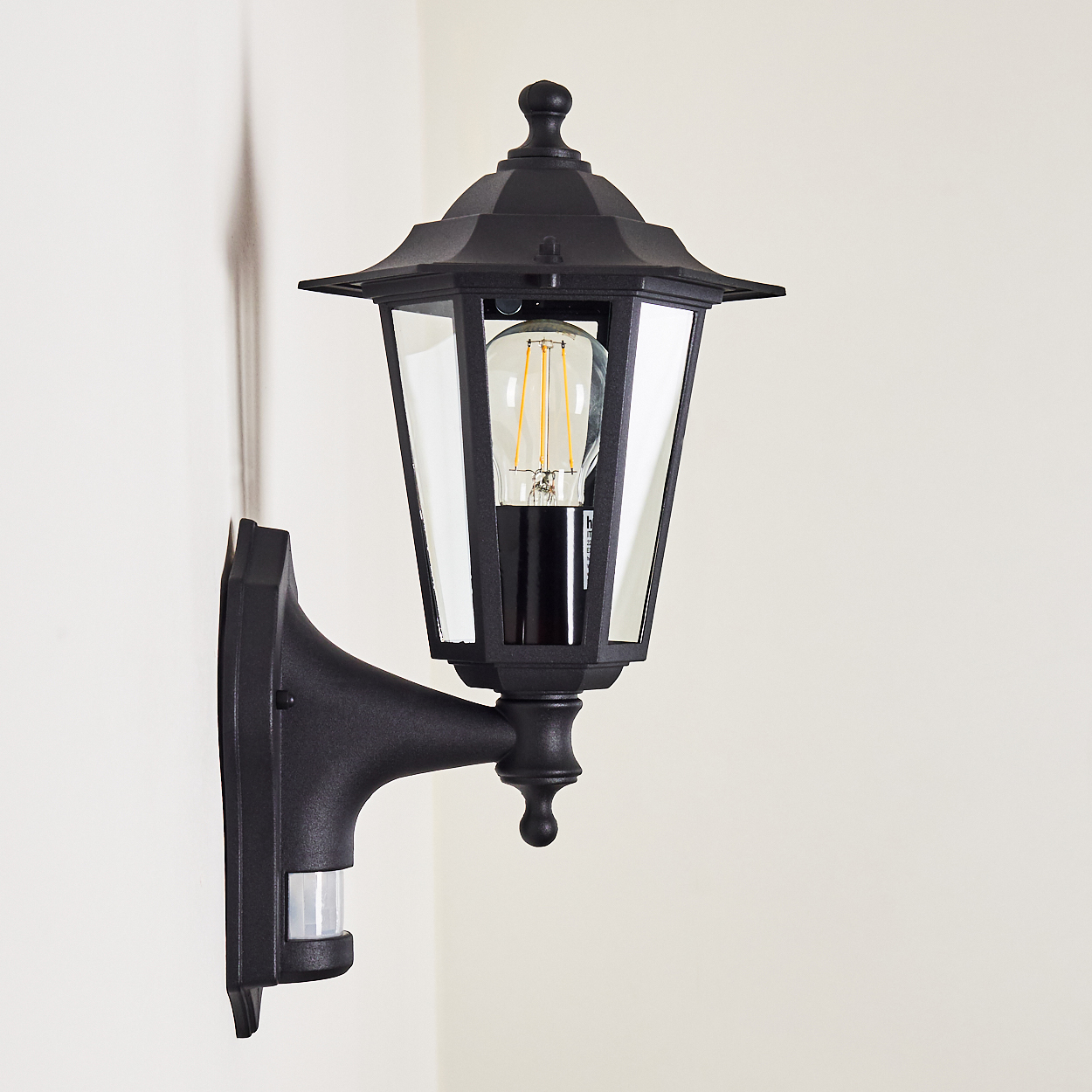 Schwarze Außenlampe mit Glasscheiben und Glühbirne, an einer weißen Wand befestigt. Die Lampe hat einen Bewegungsmelder.