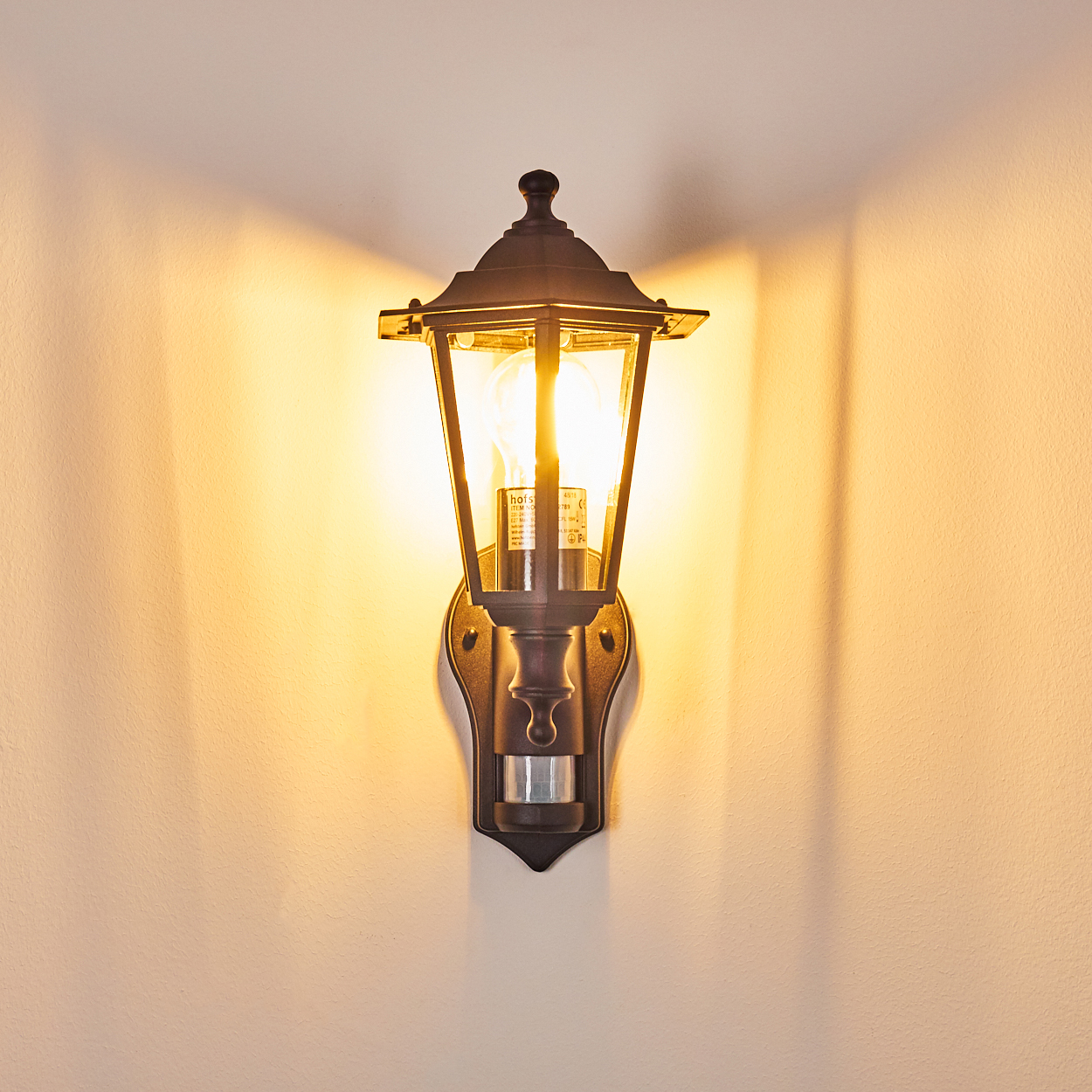 Eine dunkle Metall-Wandlampe mit einer leuchtenden Glühbirne hängt an einer beigen Wand. Das Licht wirft Schatten. Die Lampe hat einen Bewegungssensor.