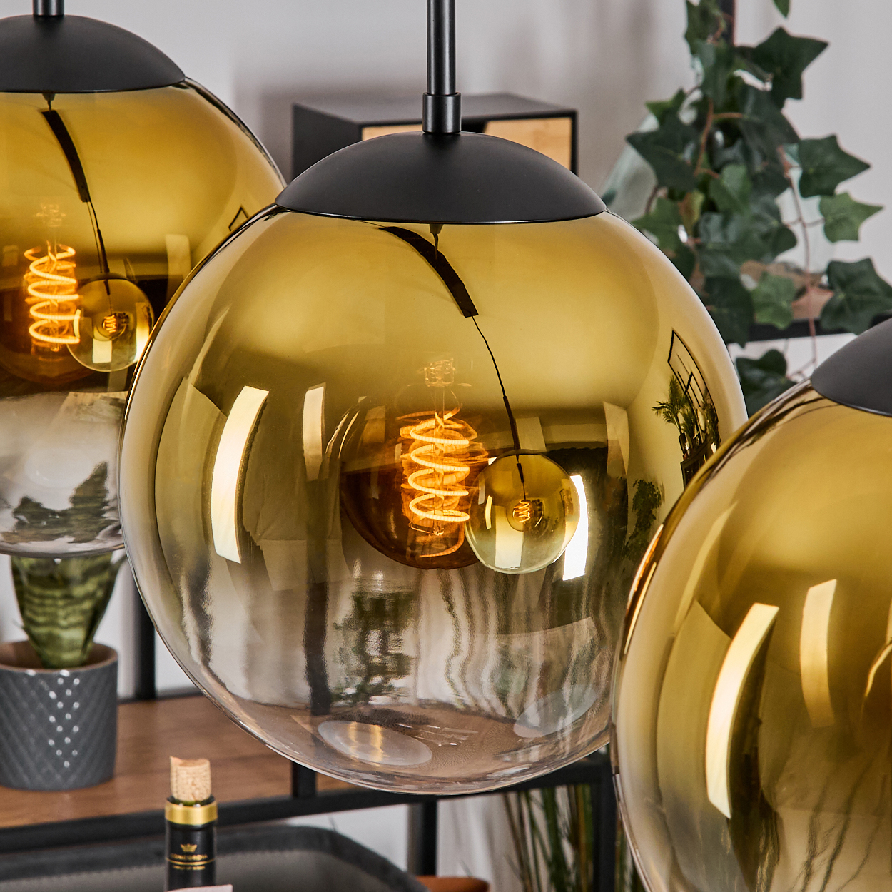 Drei goldfarbene, kugelförmige Lampen hängen von der Decke. Jede Lampe hat einen schwarzen Deckel und eine sichtbare Glühbirne.