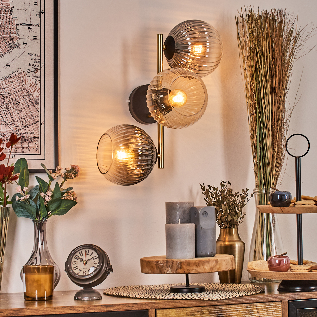 Raumdekoration mit Wandlampe, Blumen, Uhr, Kerzen und einer mehrstöckigen Ablage. Weiche Beleuchtung schafft eine gemütliche Atmosphäre.