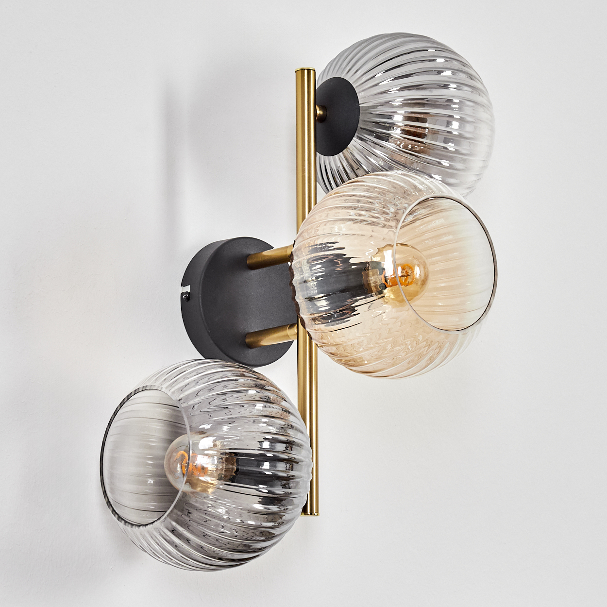 Eine Wandlampe mit drei Glaskugeln. Die Lampe hat einen goldfarbenen Rahmen und einen schwarzen Sockel. Die Kugeln sind klar und gerippt.