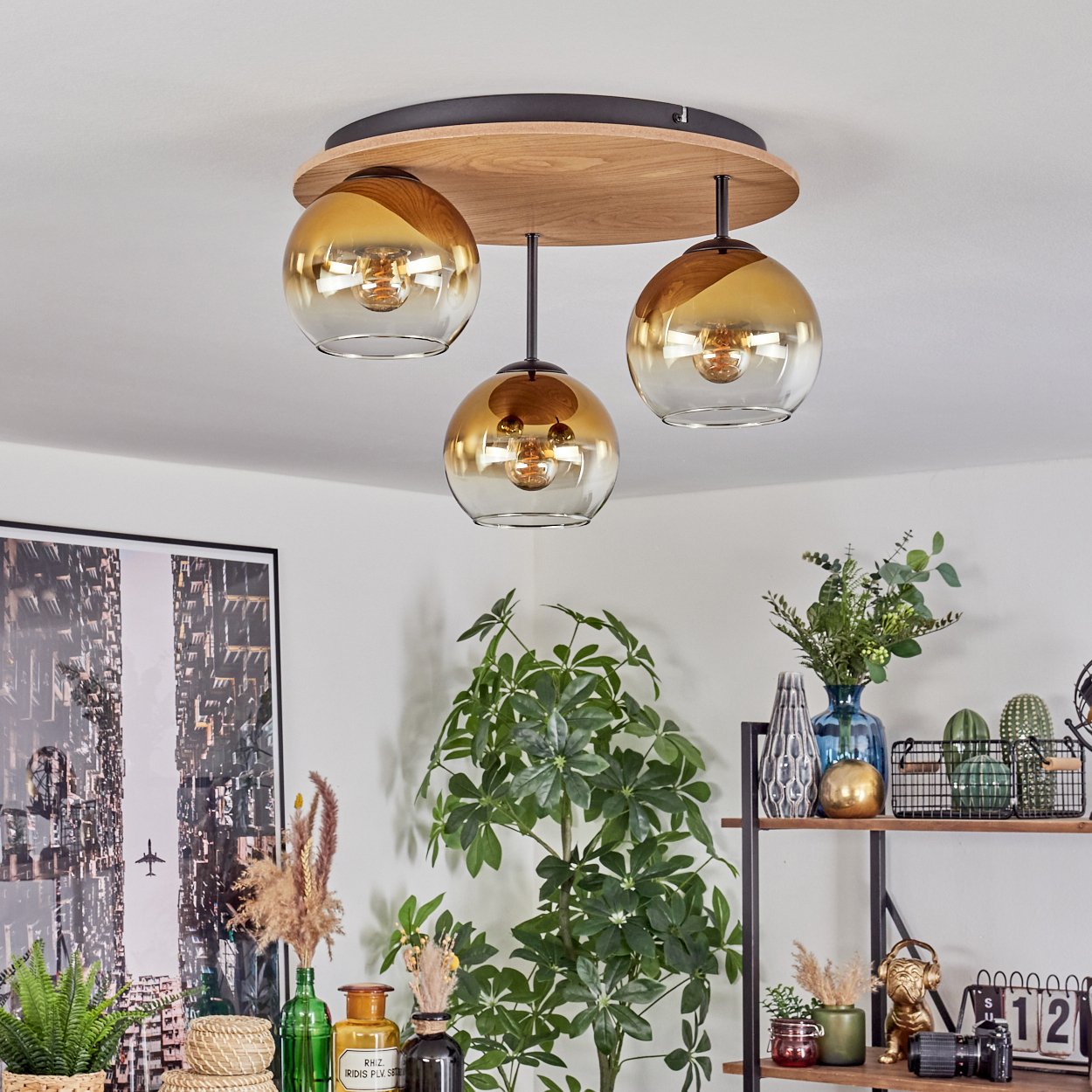 Deckenlampe mit drei goldfarbenen Glaskugeln. Hintergrund: Pflanzen, Regale mit Deko und ein Stadtbild.