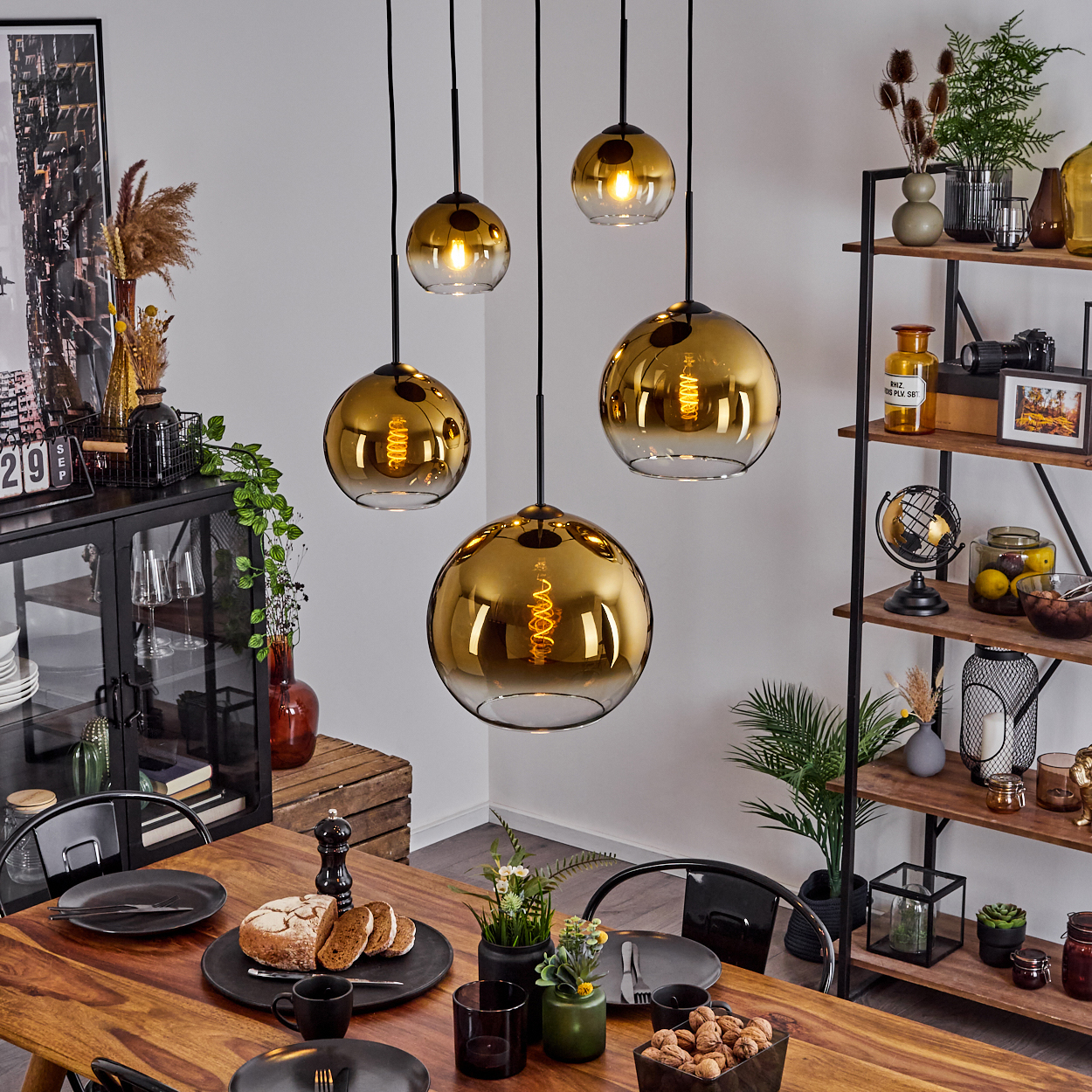 Esszimmer mit Holztisch, goldenen Pendelleuchten, Brot, Tellern und einem Regal mit Dekorationen im Hintergrund.