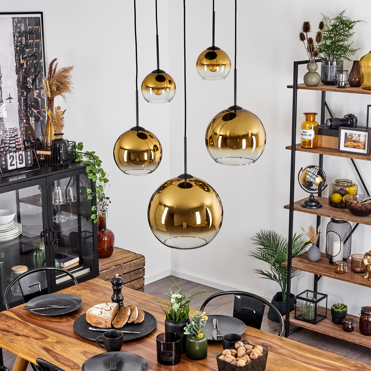 Ein Esszimmer mit Holztisch, schwarzen Stühlen und goldenen Pendelleuchten. Rechts ein Regal mit Dekorationen.