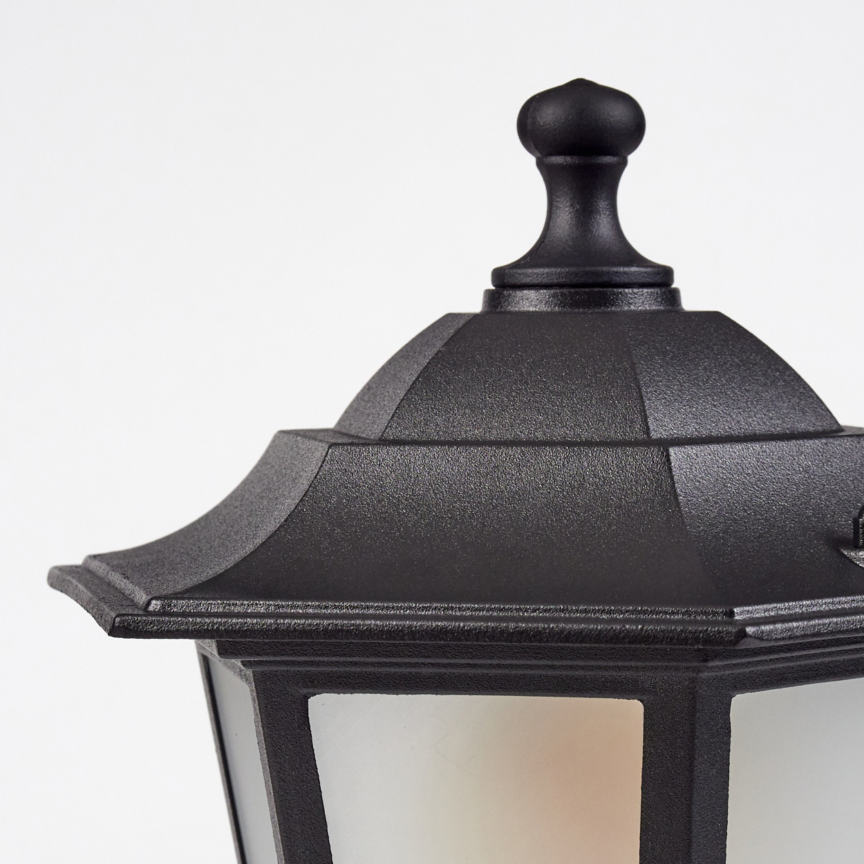 Nahaufnahme einer schwarzen Außenlampe. Die Lampe hat eine dekorative Oberseite und eine Milchglasscheibe.
