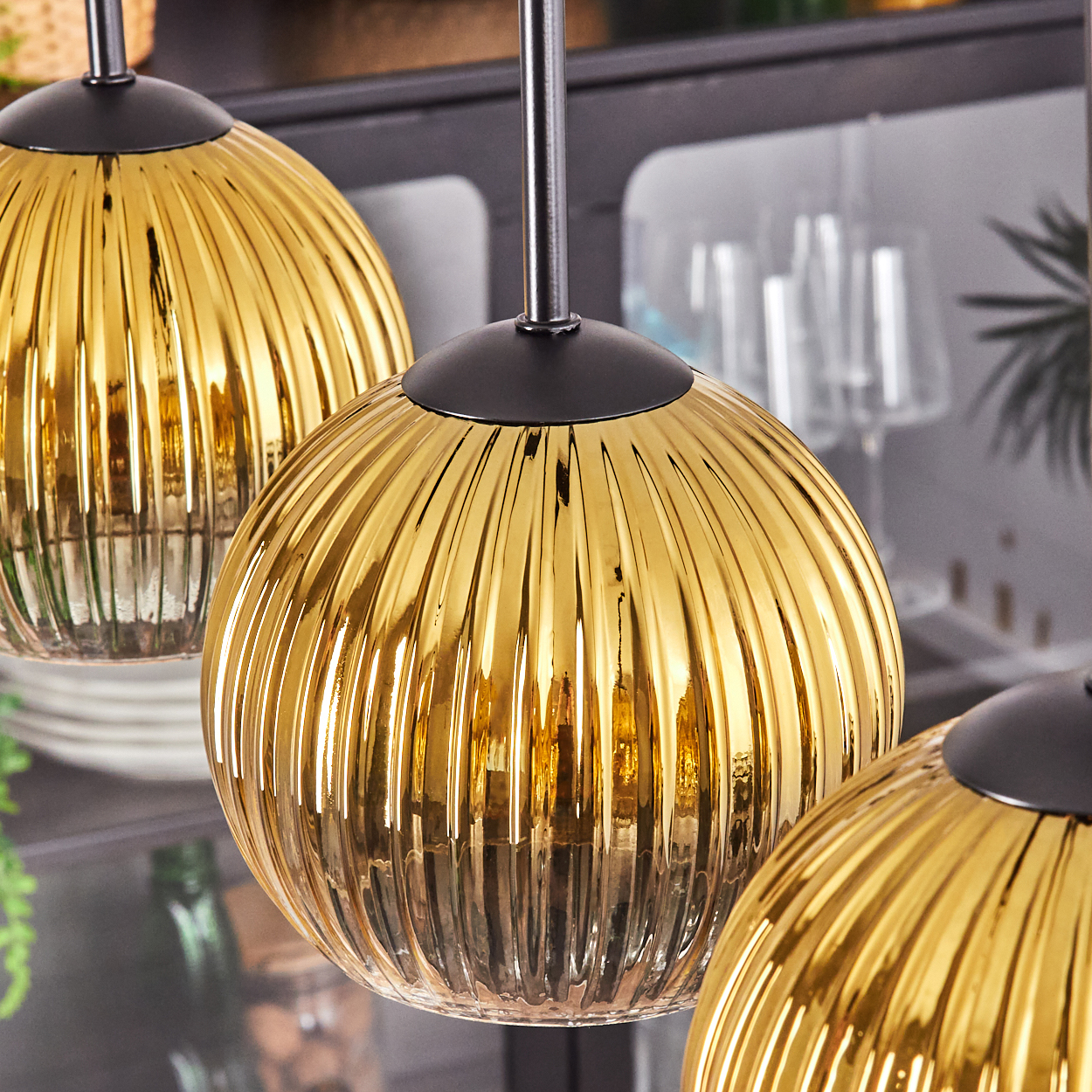 Drei goldene, geriffelte Glas-Pendelleuchten hängen an schwarzen Stangen. Der Hintergrund umfasst Küchenartikel, Teller und Gläser.