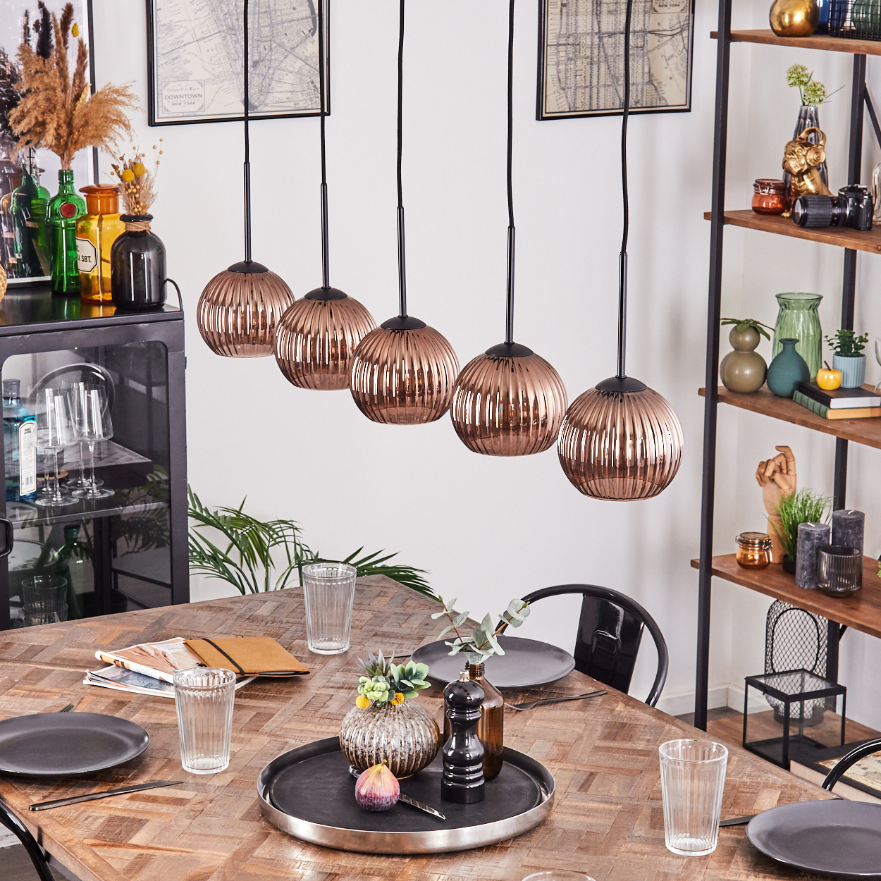 Esszimmer. Kupferfarbene Kugelleuchten hängen über einem Holztisch, der zum Abendessen gedeckt ist. Regale und gerahmte Karten befinden sich im Hintergrund.