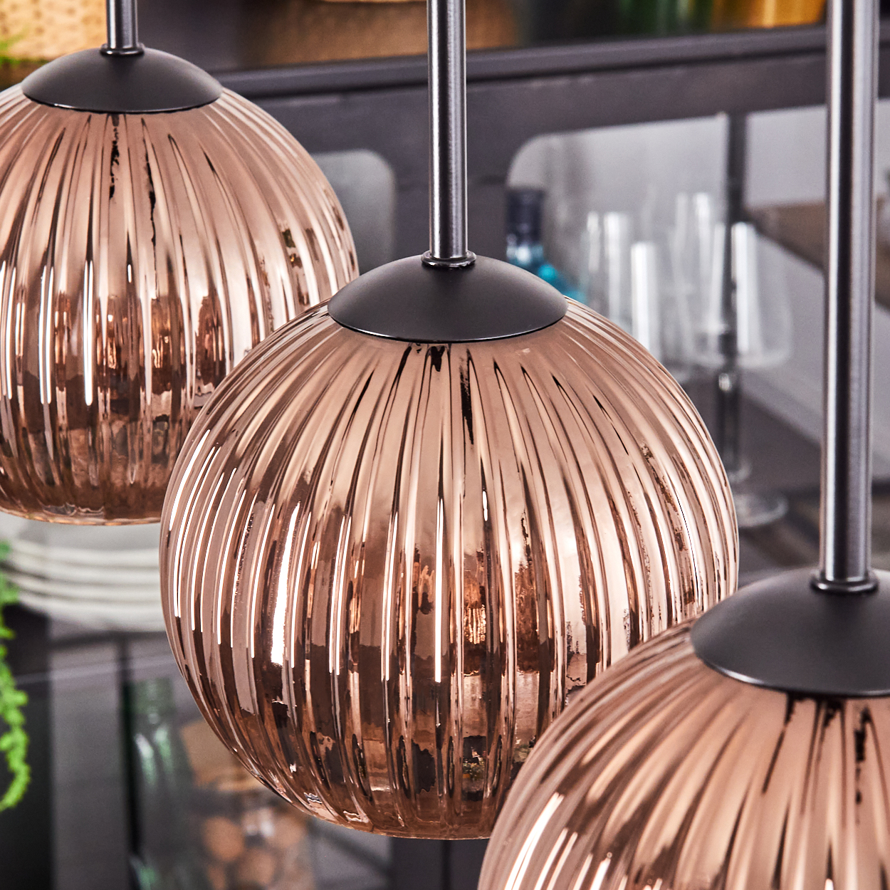 Drei kupferfarbene, gerippte Glas-Pendelleuchten hängen. Der Hintergrund zeigt einen Schrank mit Gläsern und anderen Gegenständen. Dunkle Metallstangen stützen die Leuchten.
