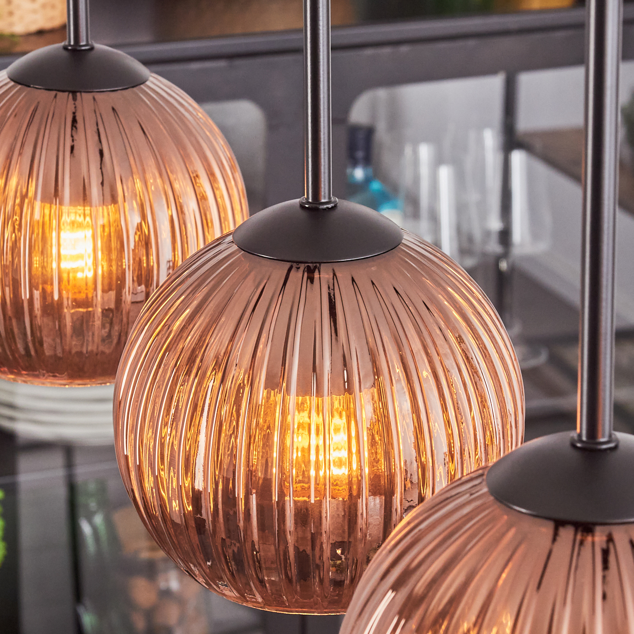 Drei kupferfarbene, gerippte Glas-Pendelleuchten mit warmem Innenlicht. Dunkle Metallstiele und -armaturen sind sichtbar.