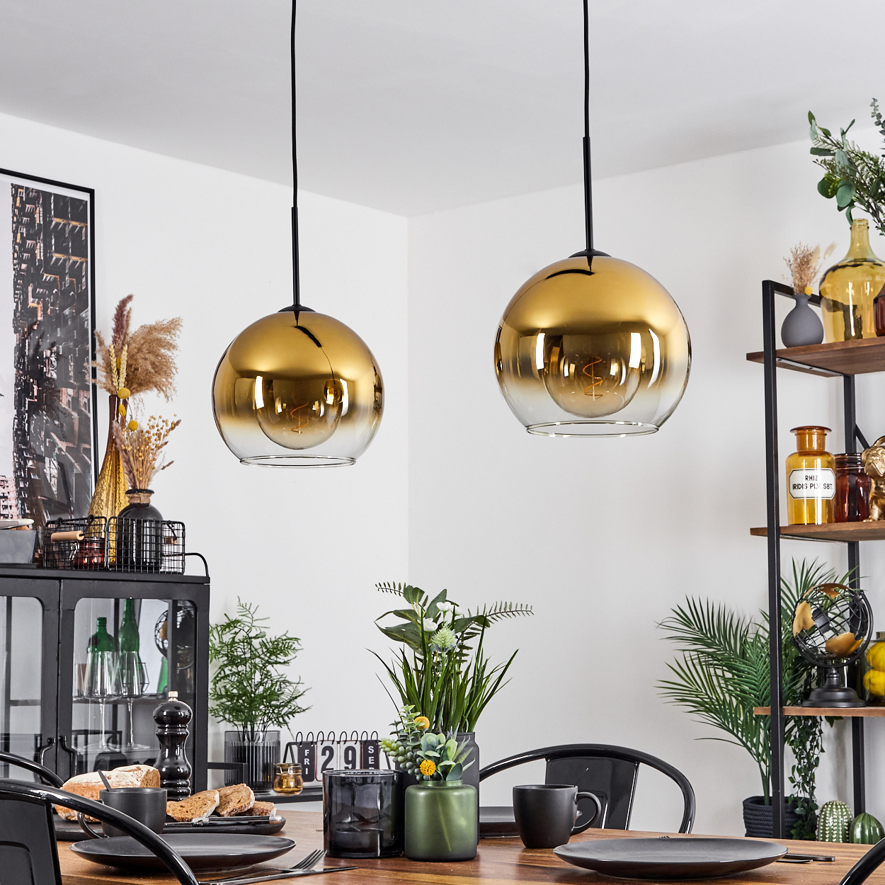 Zwei goldene Pendelleuchten hängen über einem Holzesstisch, der für eine Mahlzeit gedeckt ist. Schwarze Stühle umgeben den Tisch.
