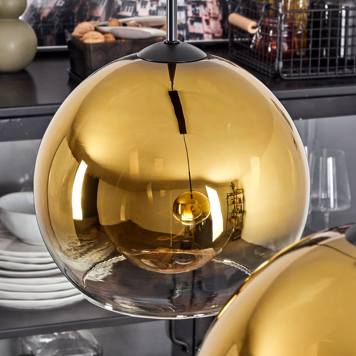 Nahaufnahme einer hängenden, runden, goldenen Glaslampe. Die Lampe reflektiert die umliegende Küche. Weitere Lampen sind im Hintergrund.