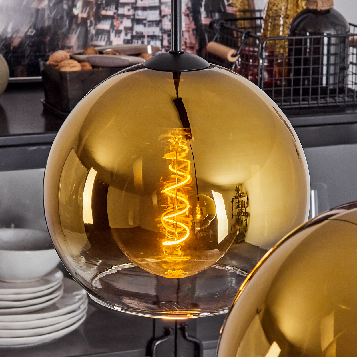 Eine goldene Glaskugel-Pendelleuchte mit sichtbarer Wendelglühbirne. Weitere Objekte sind Teller, ein schwarzer Korb und ein dunkler Hintergrund.