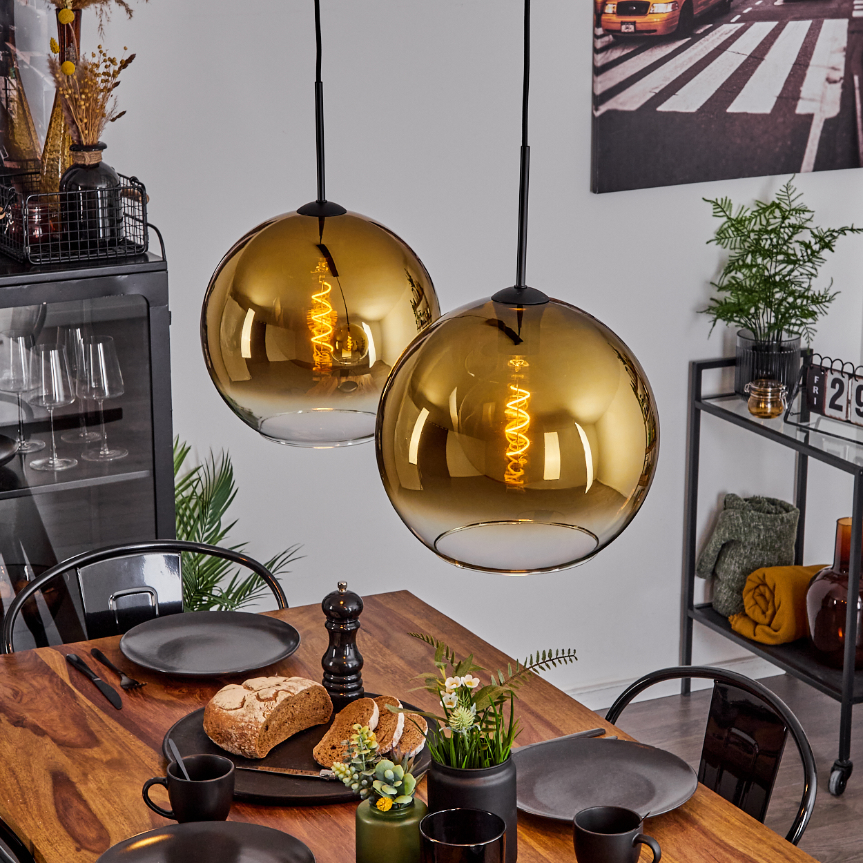 Zwei Hängelampen über einem Esstisch mit Tellern, Brot und Blumen. Hintergrund: Kunst, Pflanze und Schrank.