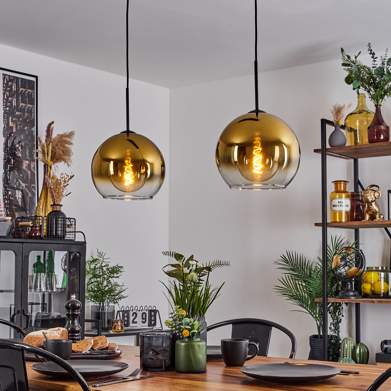Zwei goldene Pendelleuchten hängen über einem Holztisch, der für eine Mahlzeit gedeckt ist. Regale im Hintergrund halten Dekorationen.