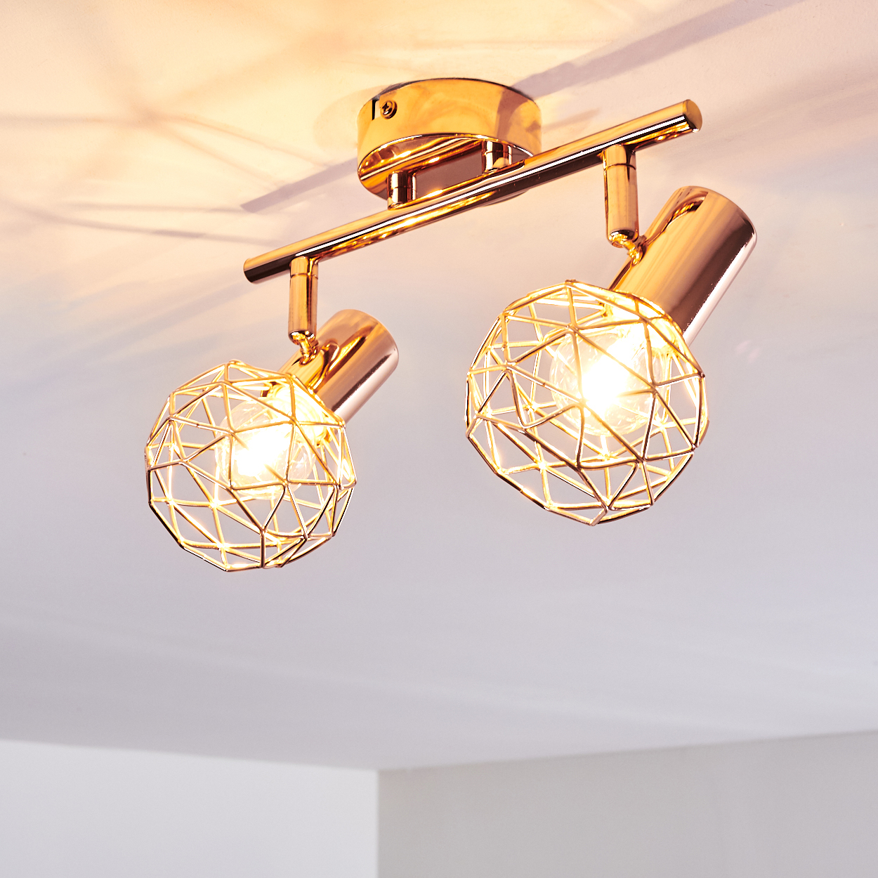 Eine goldene Deckenleuchte mit zwei kugelförmigen, käfigartigen Schirmen. Die Lichter sind an und beleuchten die Leuchte und die Decke.
