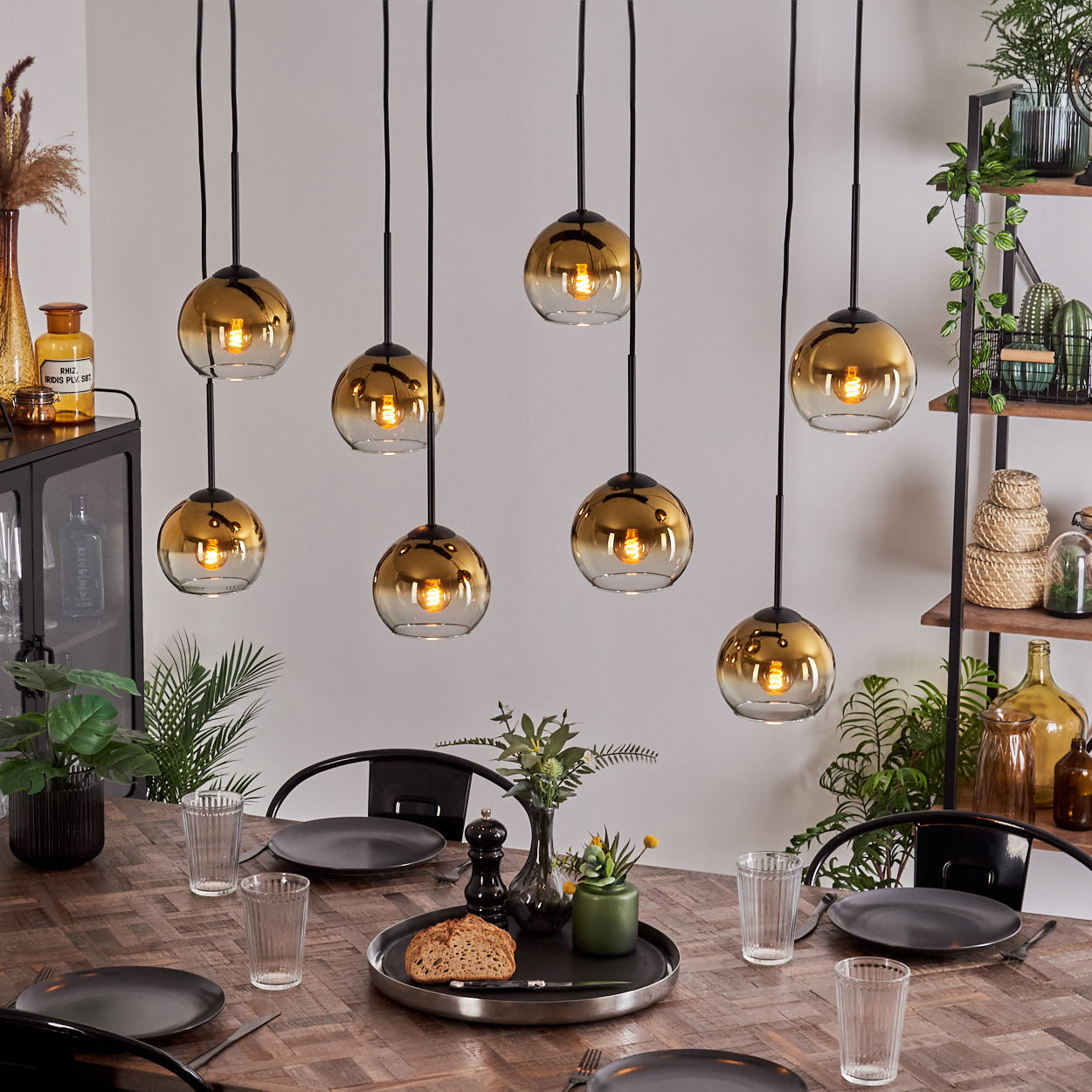 Esstisch mit Tellern, Gläsern und Brot. Oben acht goldene Pendelleuchten. Pflanzen und Regale im Hintergrund.