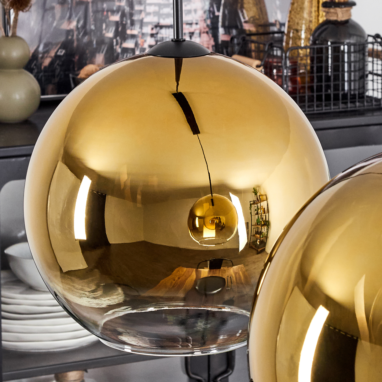 Zwei goldene Glas-Pendelleuchten. Die Lichter reflektieren den Raum, mit Regalen, Vasen und Geschirr im Hintergrund sichtbar.
