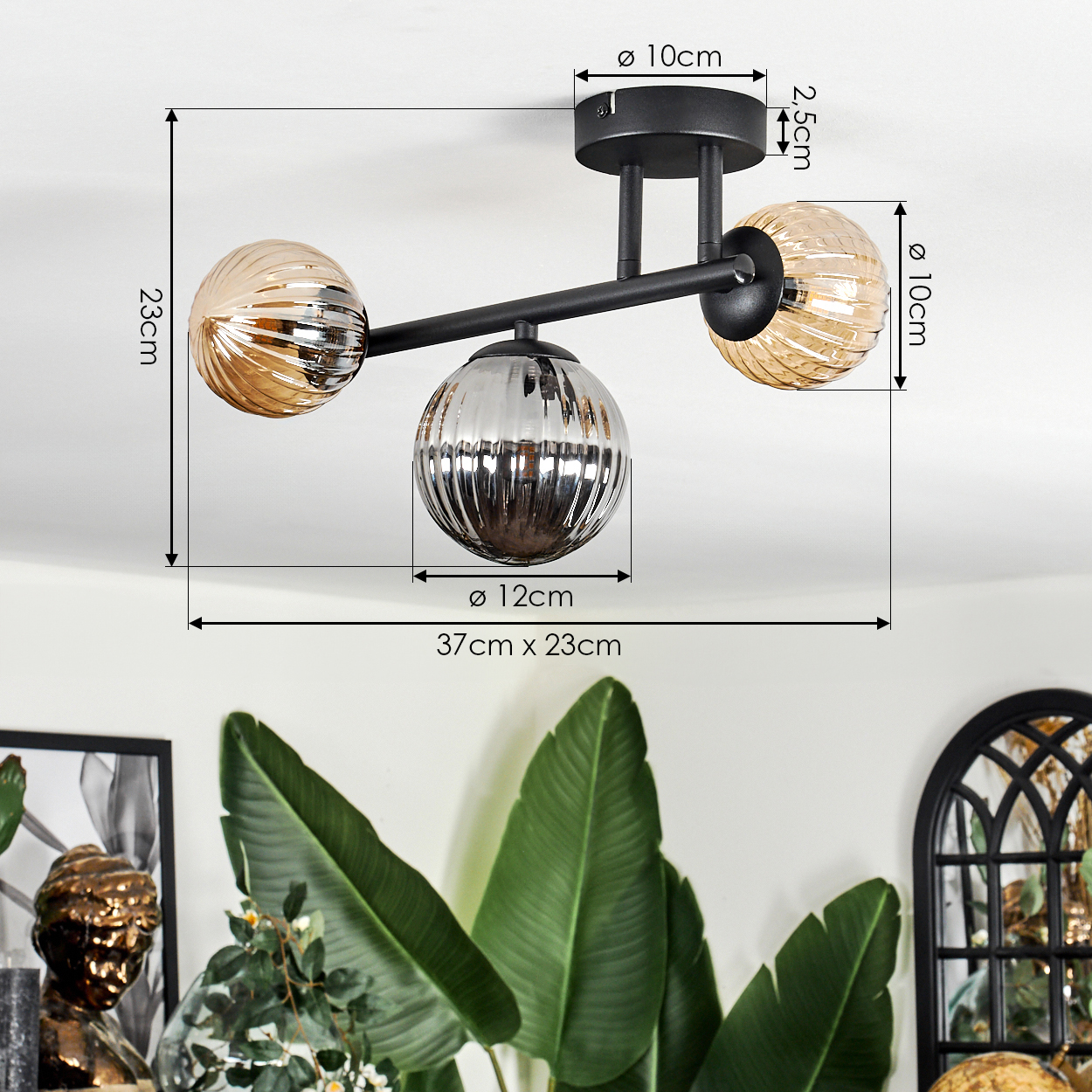 Eine moderne Deckenlampe mit drei Glaskugeln. Die Lampe ist schwarz, die Kugeln sind bernsteinfarben und silber. Grüne Pflanzen im Vordergrund.