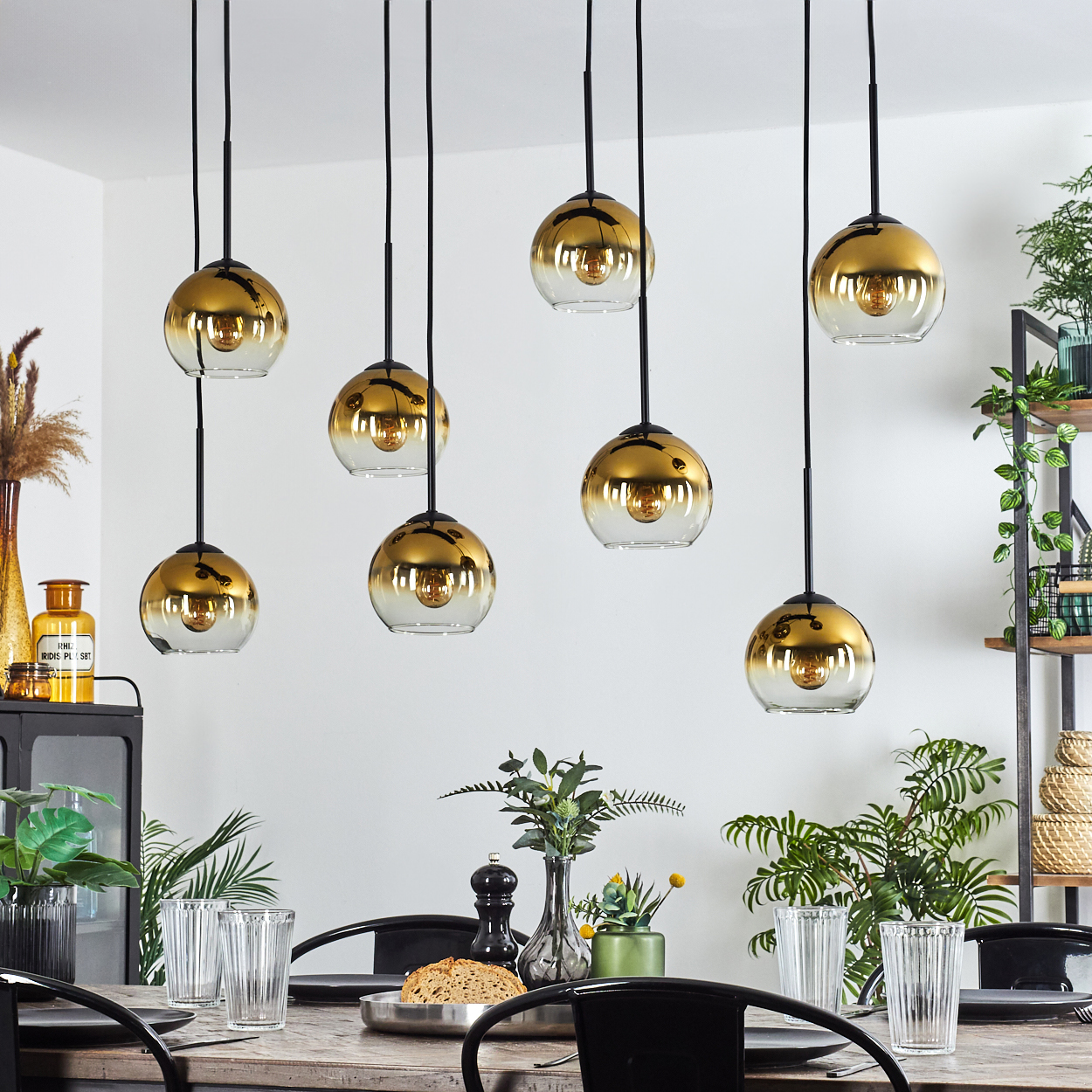 Esszimmer mit goldenen Pendelleuchten. Tisch mit Gläsern und Tellern, Pflanzen im Hintergrund. Weiße Wand.