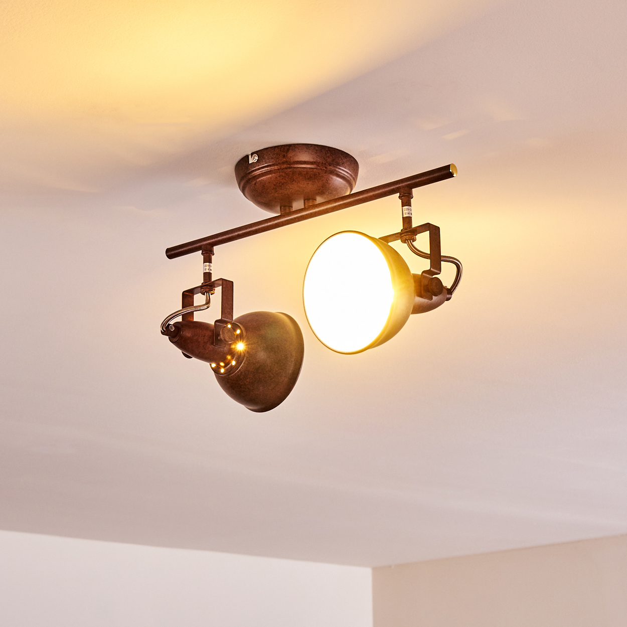 Deckenstrahler mit zwei verstellbaren Köpfen. Er hat ein braunes, industrielles Design und wirft warmes Licht. Die Decke ist weiß.