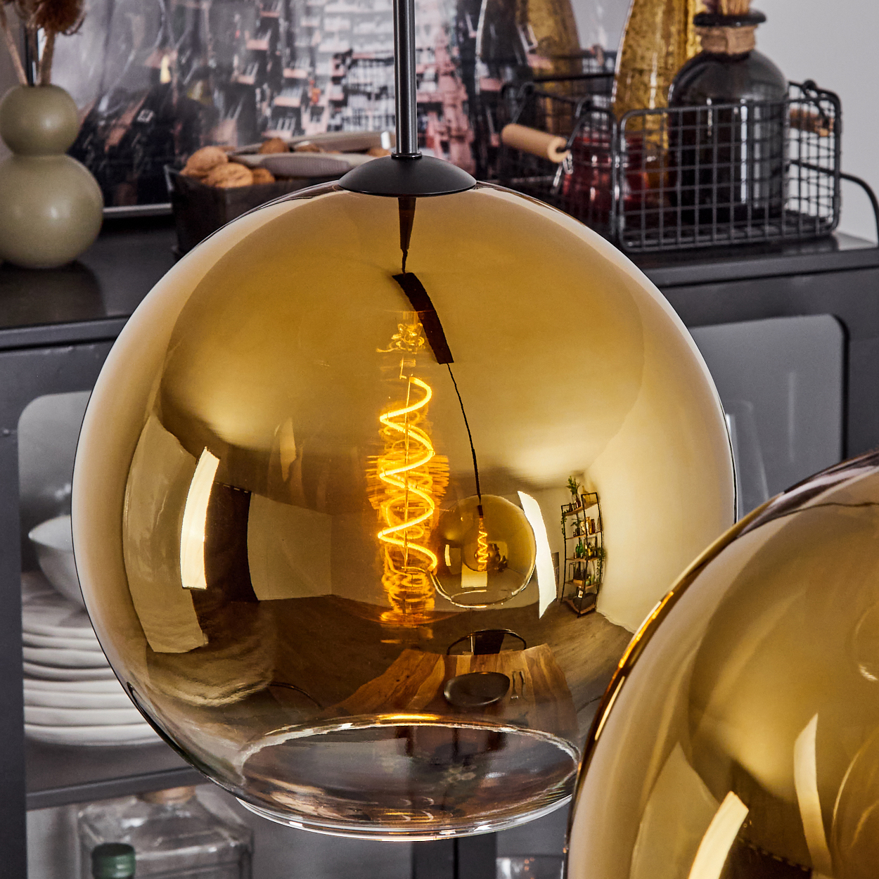 Nahaufnahme einer goldenen Glas-Pendelleuchte mit einer Wendelglühbirne. Andere Lampen und Dekorationen sind im Hintergrund.