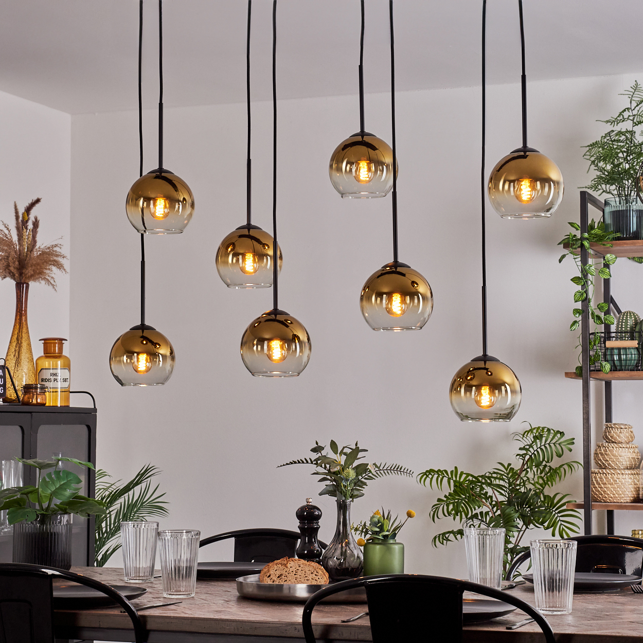 Esszimmer mit goldenen Hängeleuchten. Tisch mit Gläsern und Brot gedeckt. Pflanzen und Regale im Hintergrund.