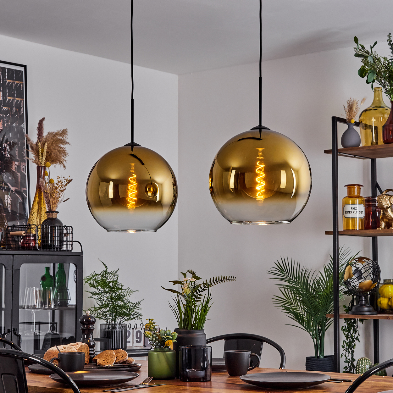 Zwei goldene Pendelleuchten hängen über einem Esstisch, der mit Tellern, Brot und Pflanzen gedeckt ist. Regale zeigen Vasen und Dekoration.