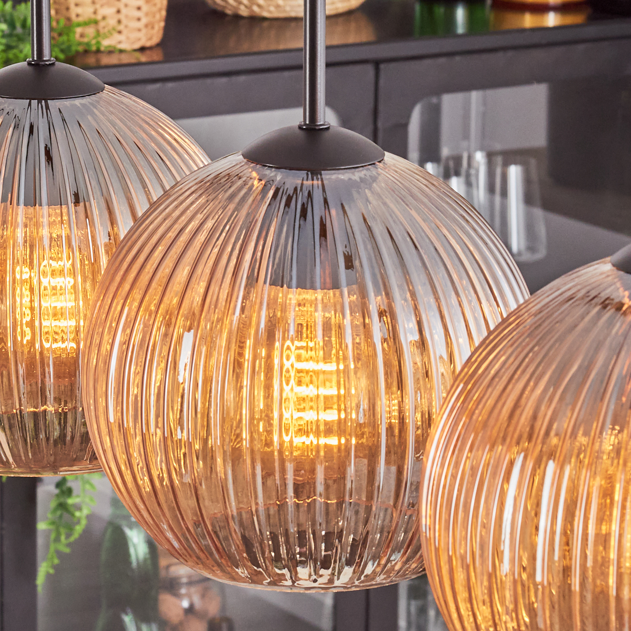Drei orangefarbene Glas-Pendelleuchten mit sichtbaren Glühbirnen. Sie hängen vor einem dunklen Hintergrund mit etwas Grün.