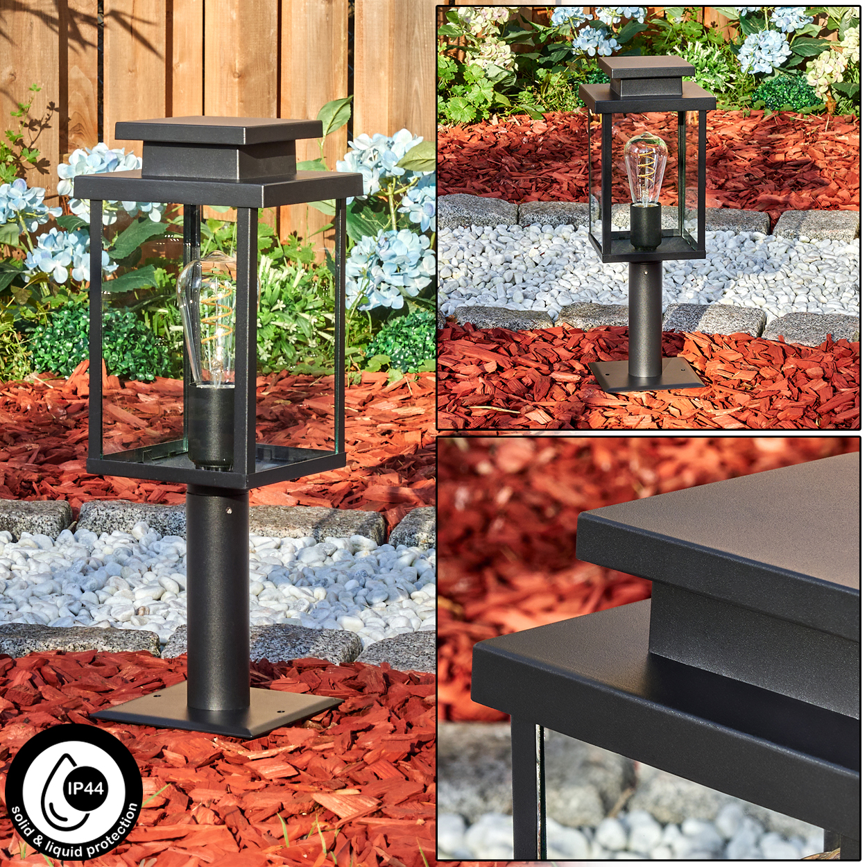 Eine schwarze Außenlampe mit Glühbirne steht auf roter Mulch. Im Hintergrund sind weiße Steine, Blumen und ein Holzzaun zu sehen.