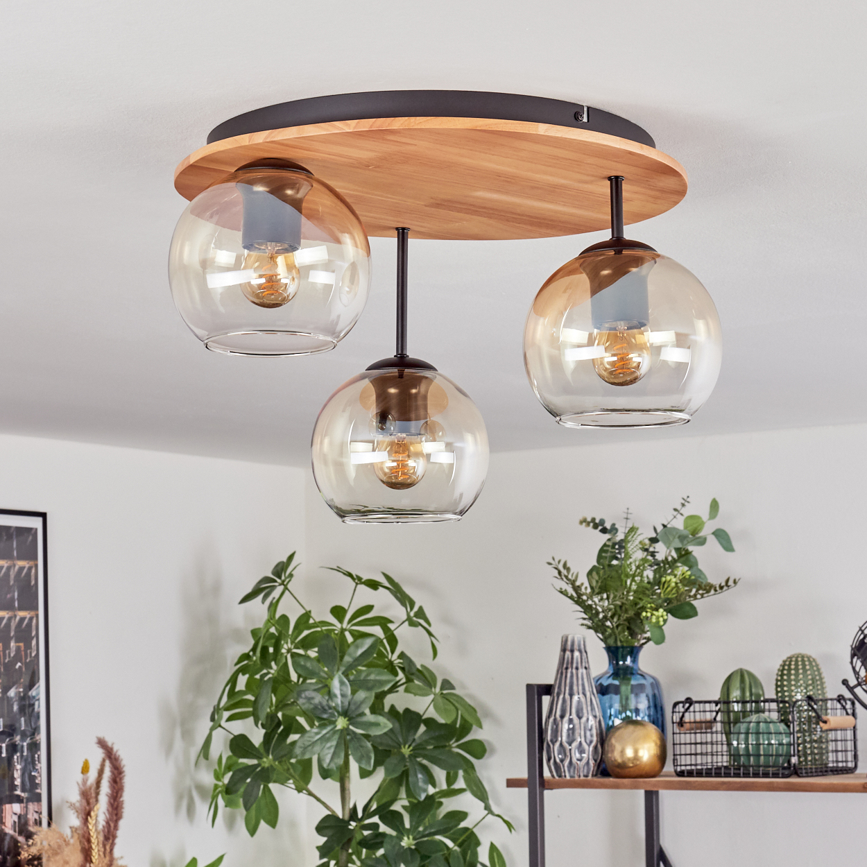 Eine moderne Deckenlampe mit drei hängenden Glaskugeln. Die Basis ist aus Holz und schwarzem Metall. Pflanzen und Dekoration im Hintergrund.