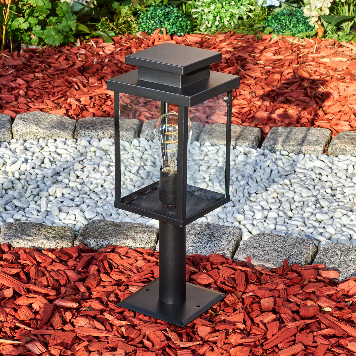 Schwarze Außenlaterne mit sichtbarer Glühbirne, in einem Garten mit roter Mulche, weißen Steinen und grünen Pflanzen.