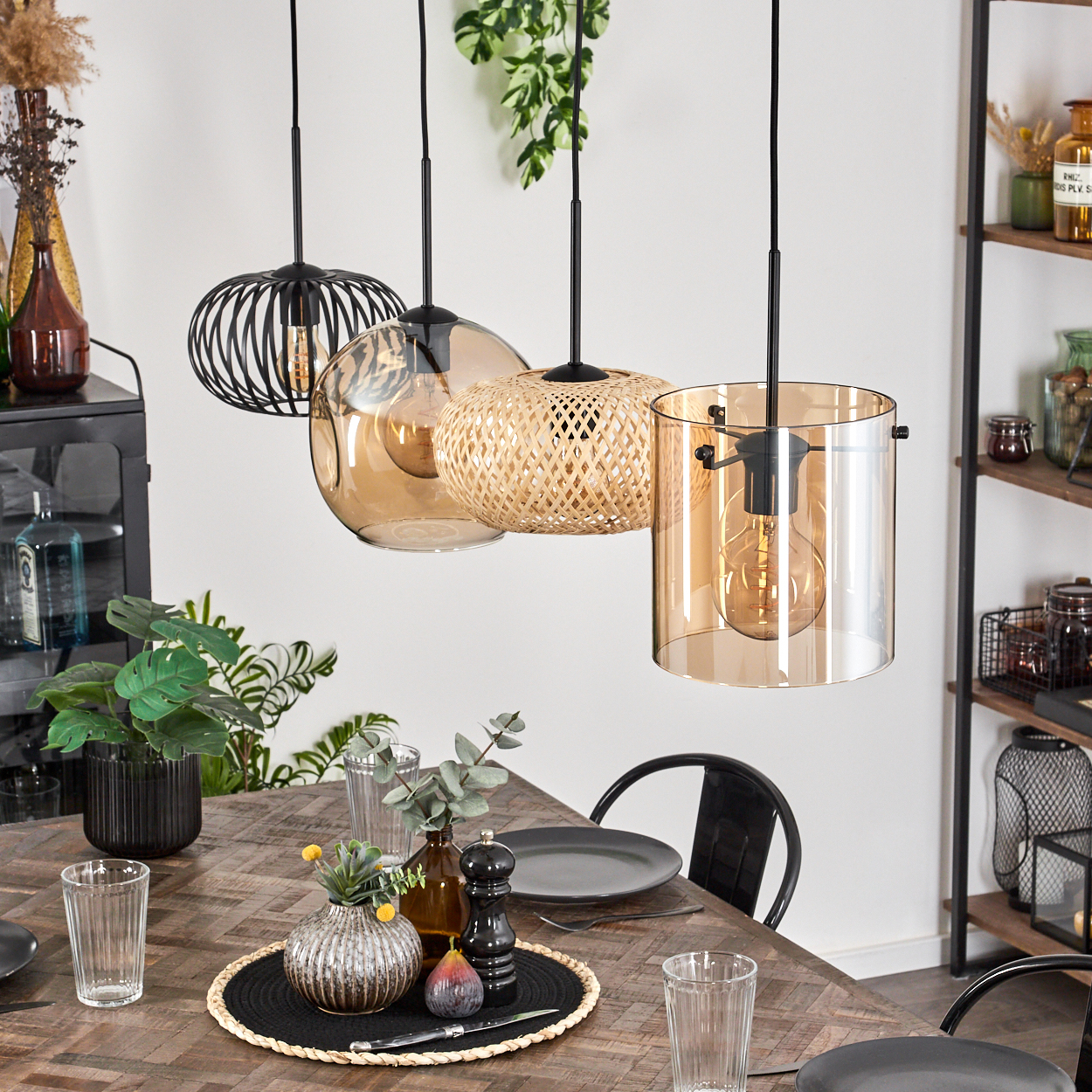 Esszimmer mit Holztisch. Vier Hängelampen. Tisch gedeckt mit Gläsern, Tellern, einer Vase und einer Feige. Regale mit Gläsern.