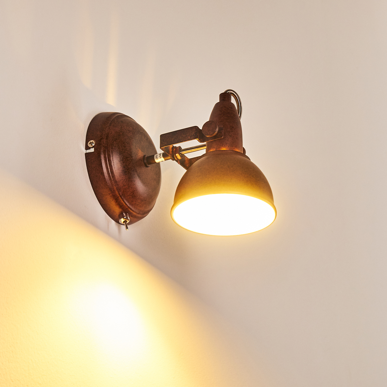 Eine rostbraune Wandlampe mit rundem Sockel und fokussiertem Licht. Das Licht ist an und wirft einen warmen Schein auf die Wand.