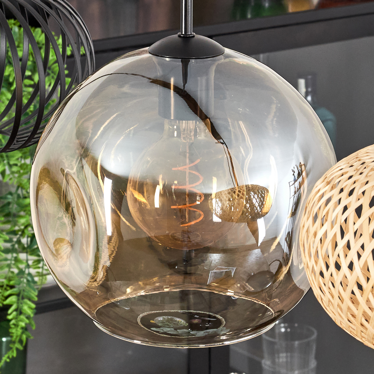 Eine klare Glaskugel-Pendelleuchte mit sichtbarer Glühbirne. Ein schwarzer Metallkäfig und eine gewebte Lampe sind ebenfalls zu sehen.
