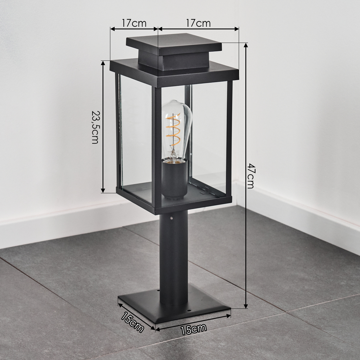 Schwarze Außenlampe mit Glasscheiben und einer Glühbirne. Sie steht auf quadratischer Basis auf Fliesen.