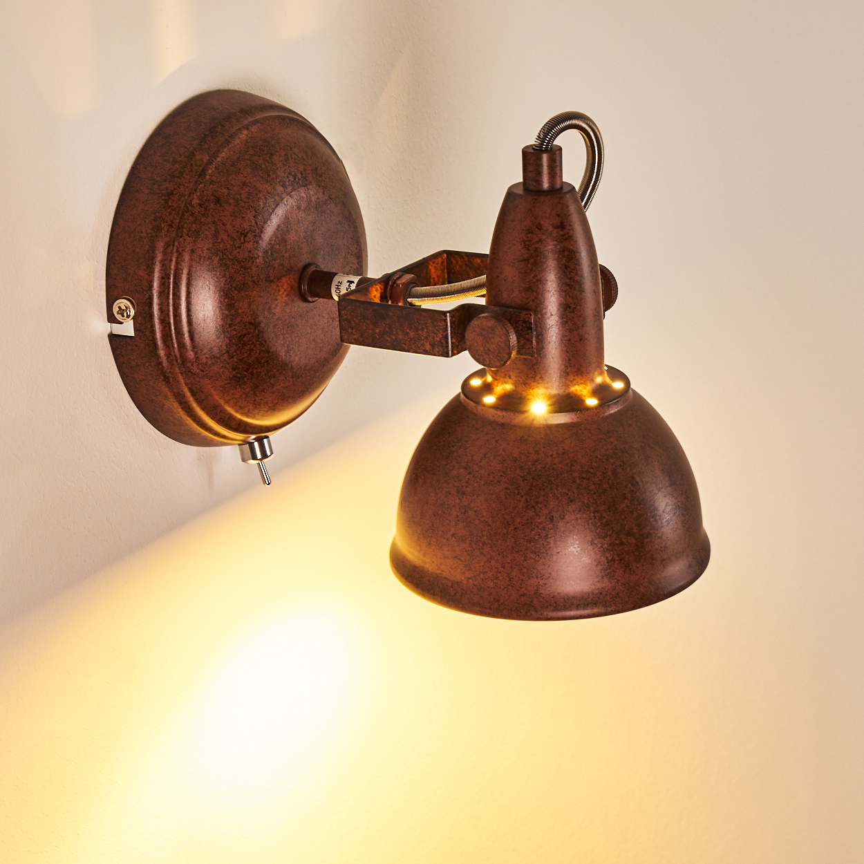 Eine rostbraune Wandlampe mit gewölbtem Schirm und Schalter. Das Licht beleuchtet die Wand unter der Lampe.