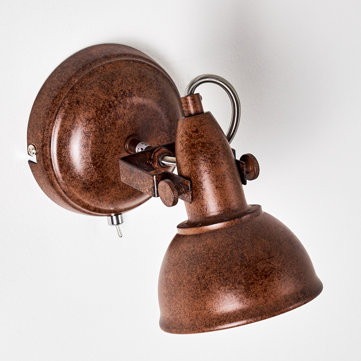 Ein bronzefarbener, an der Wand befestigter Strahler. Er hat eine runde Basis, einen flexiblen Arm und einen verstellbaren Lampenschirm, vor einer weißen Wand.