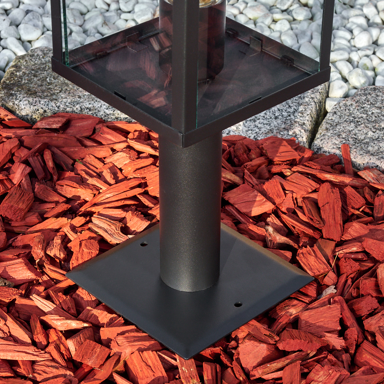 Eine schwarze Metalllaterne auf roter Mulch, mit Glasplatte. Weiße und graue Steine befinden sich im Hintergrund.