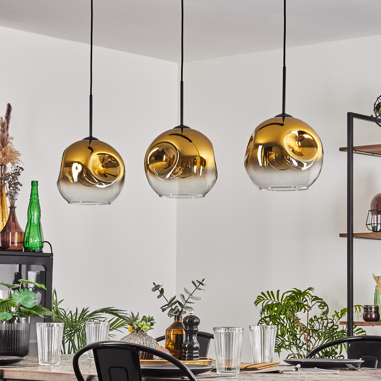 Drei gold- und klarglasige Pendelleuchten hängen über einem Esstisch mit Gläsern und Pflanzen. Ein Regal und Dekoration sind im Hintergrund.