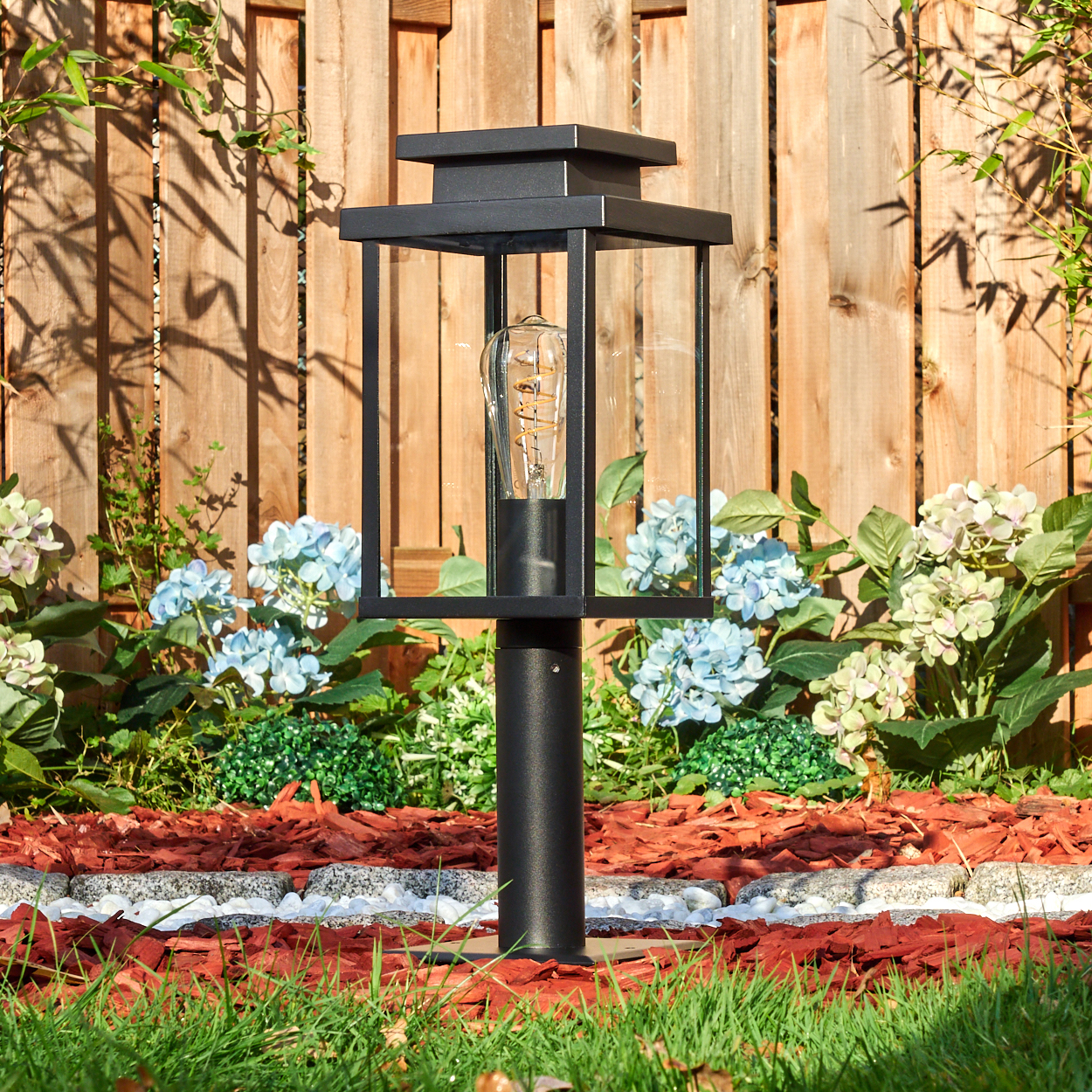 Eine schwarze Außenlampe steht in einem Garten. Die Lampe hat eine Glühbirne. Dahinter ist ein Holzzaun und Blumen.