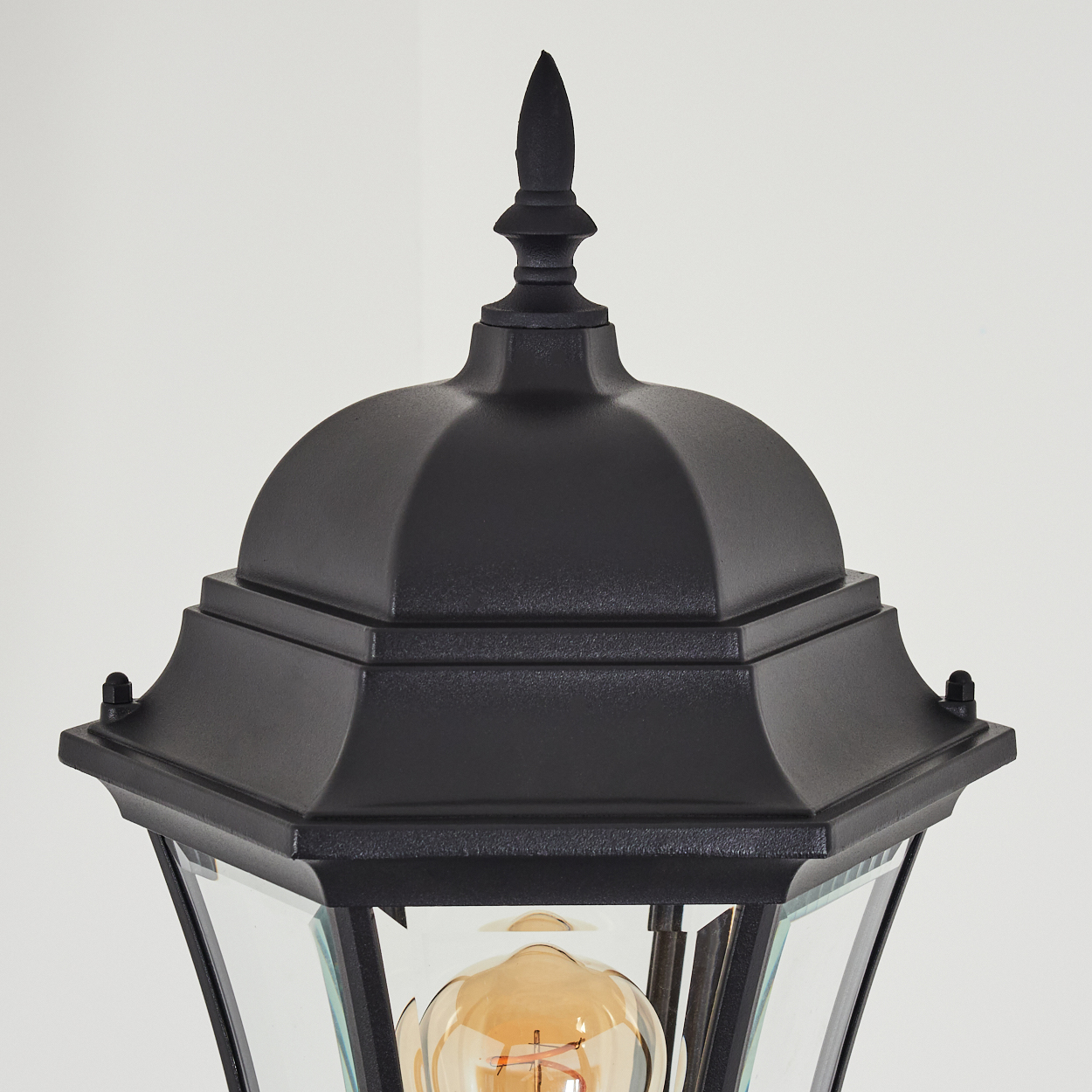 Nahaufnahme einer schwarzen Außenlampe. Sie hat eine spitze Oberseite, ein gewölbtes Dach und Glasscheiben. Die Glühbirne ist sichtbar. Weißer Hintergrund.