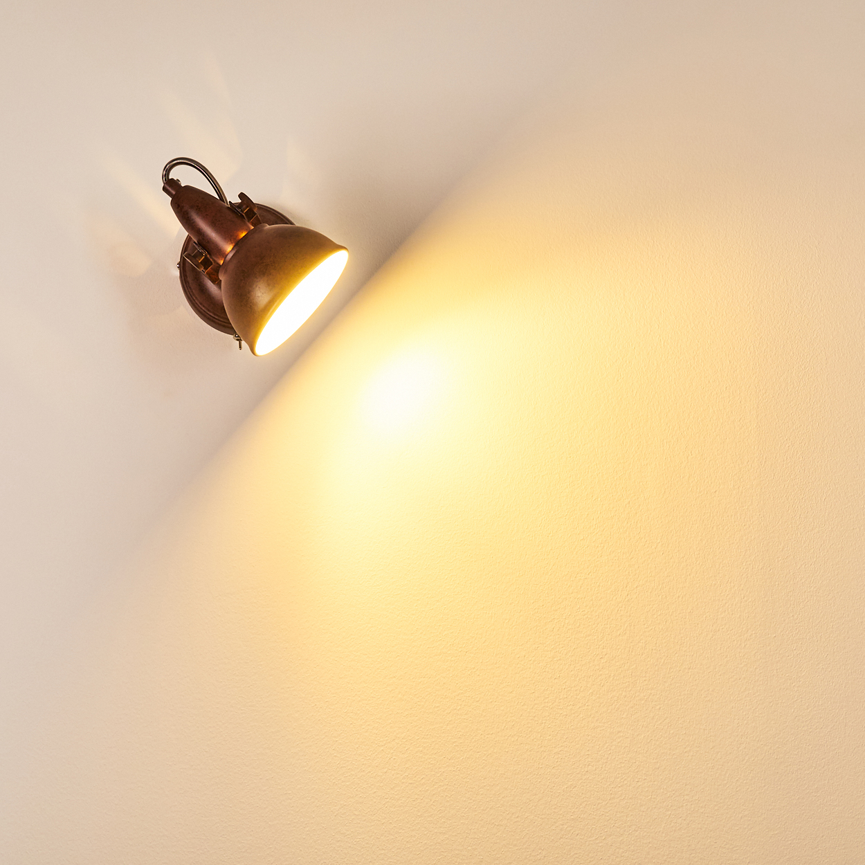 Ein bronzefarbener Scheinwerfer an einer weißen Wand. Das Licht scheint hell und beleuchtet einen großen Bereich der Wand.
