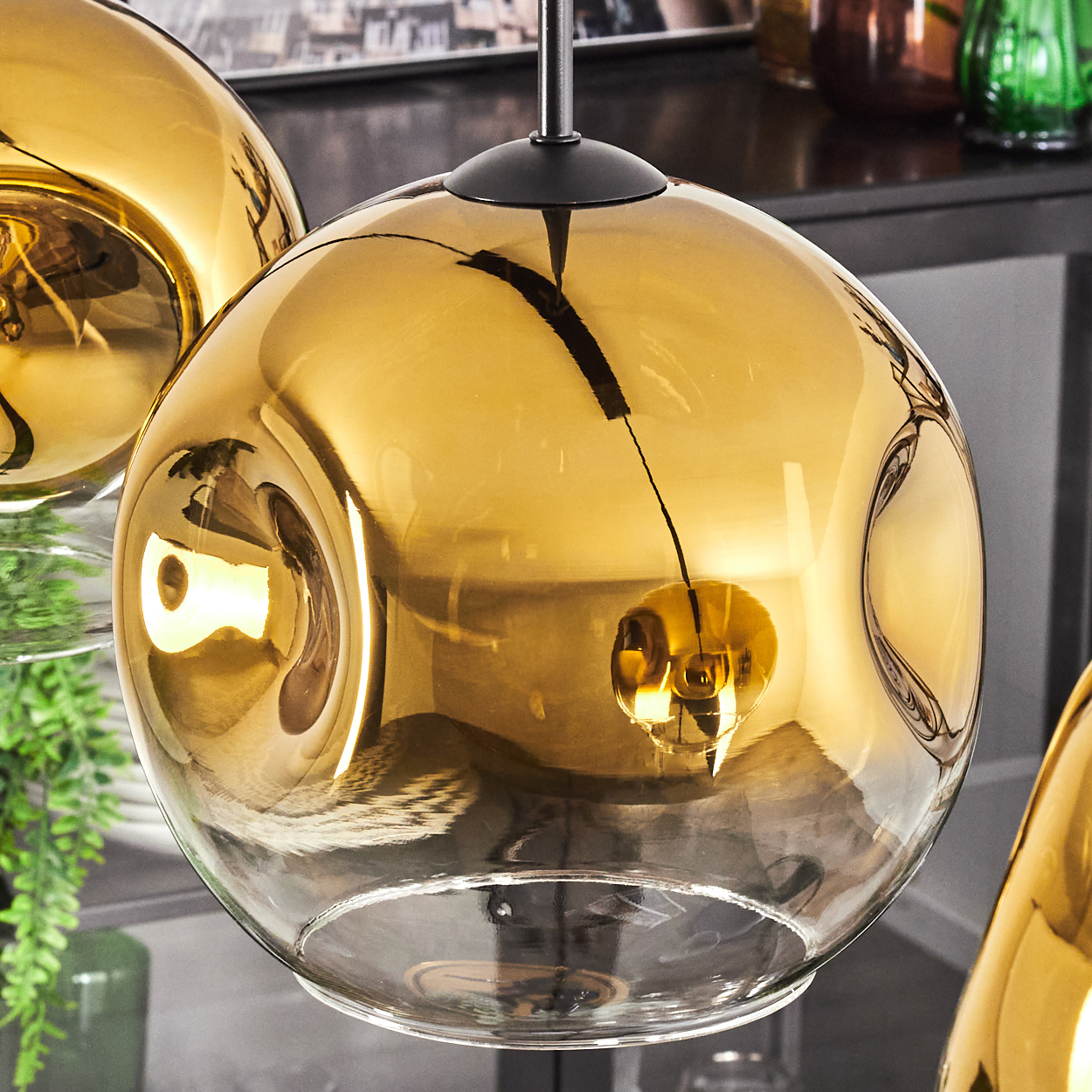 Nahaufnahme einer goldenen Glaskugel-Pendelleuchte. Weitere Lampen sind sichtbar, zusammen mit Pflanzen und Regalen im Hintergrund.