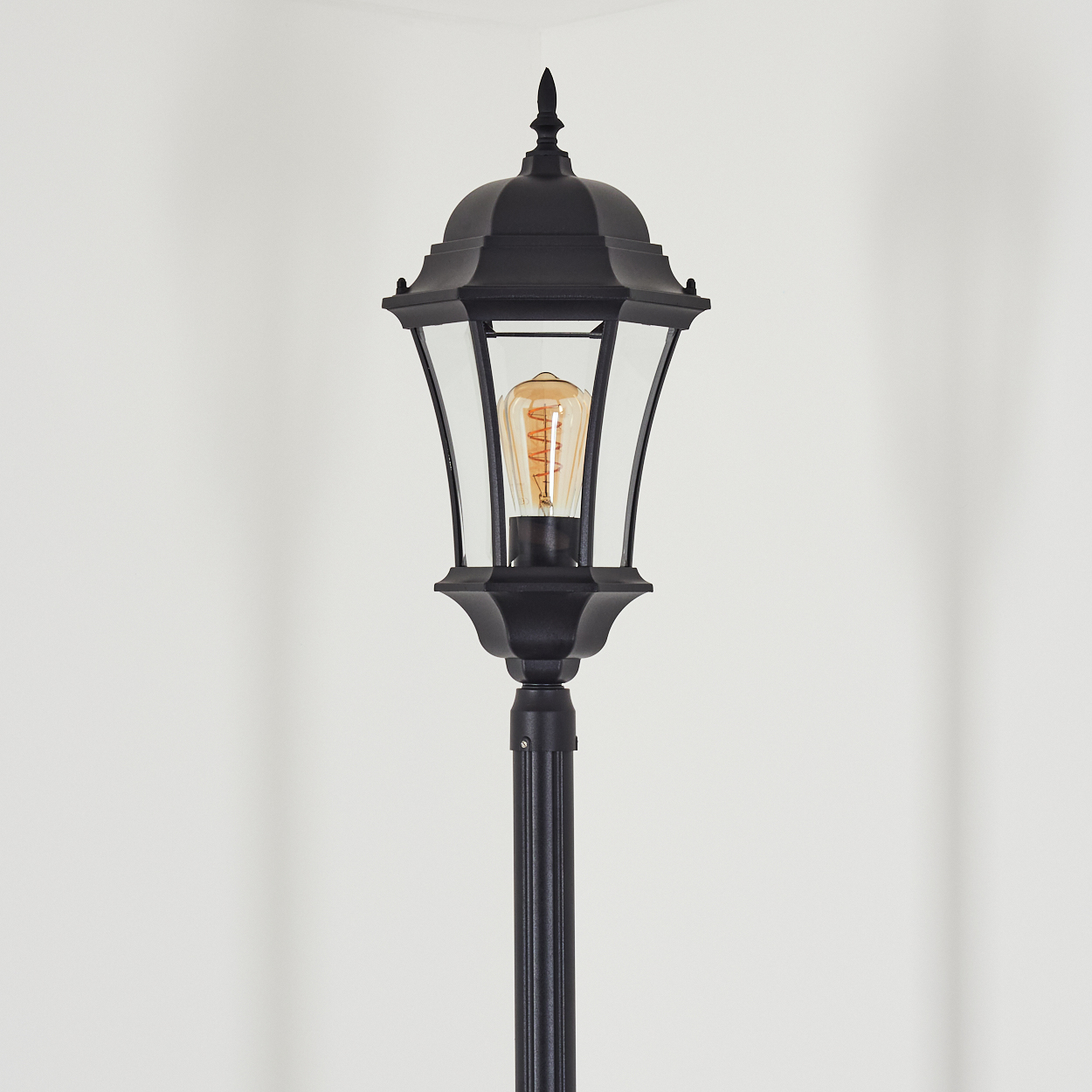 Schwarze Außenlampe mit Glasgehäuse und dekorativer Spitze, mit einer warm getönten Glühbirne, vor weißem Hintergrund.