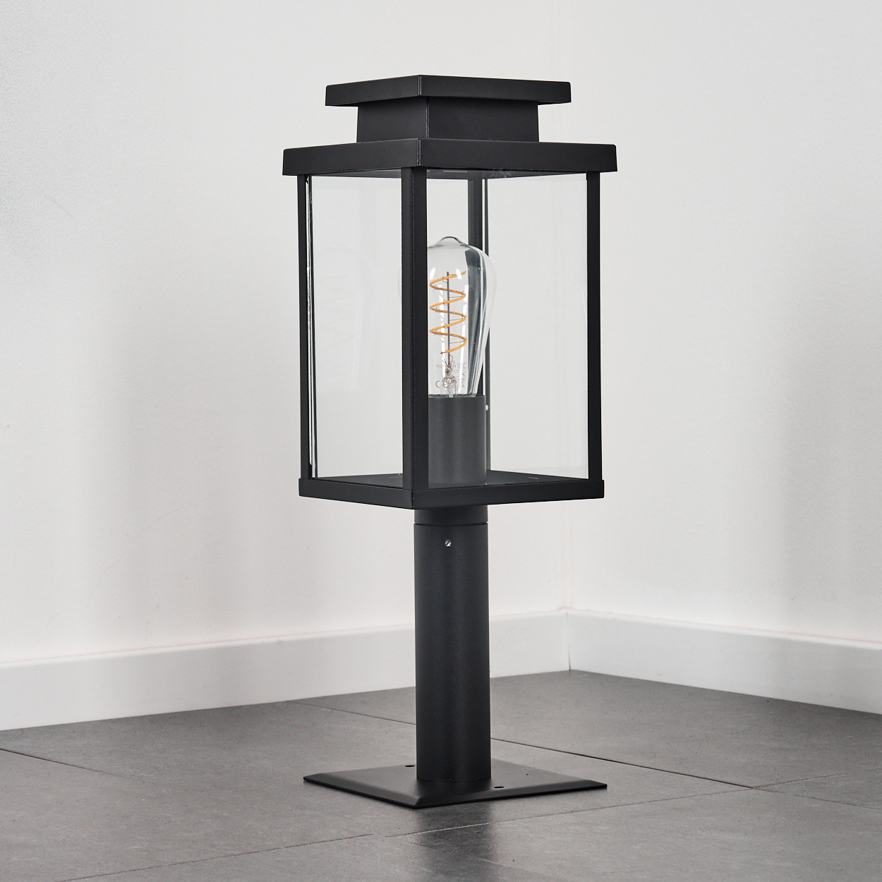 Eine schwarze Außenlampe steht auf einem grauen Fliesenboden. Sie hat ein quadratisches Glasgehäuse mit einer Glühbirne. Weiße Wand im Hintergrund.