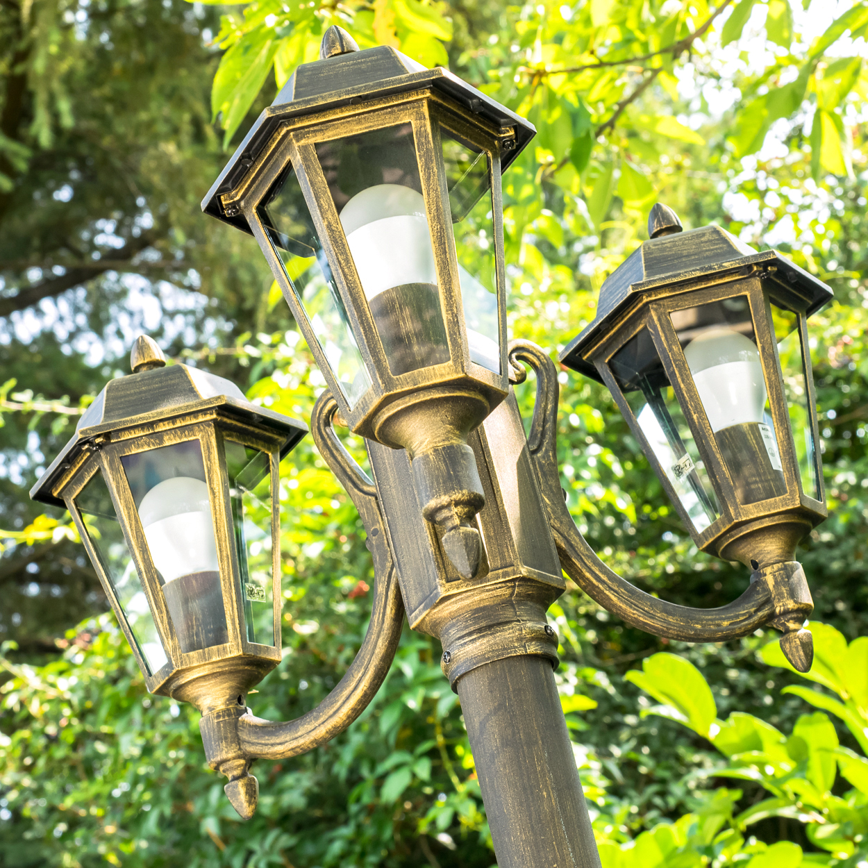Eine verzierte bronzefarbene Straßenlaterne mit drei Lampen, jede mit einer weißen Glühbirne, vor einem Hintergrund aus grünen Blättern.