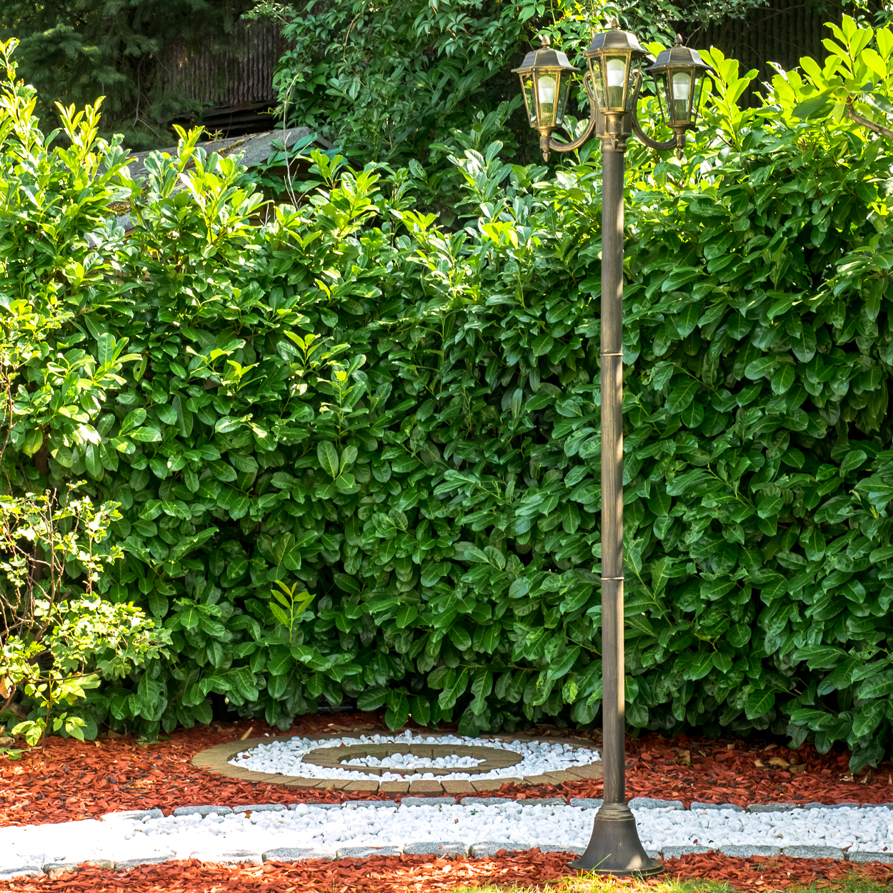 Eine bronzefarbene Laterne steht vor einer dichten grünen Hecke. Weiße Steine und roter Mulch bilden einen Weg im Vordergrund.