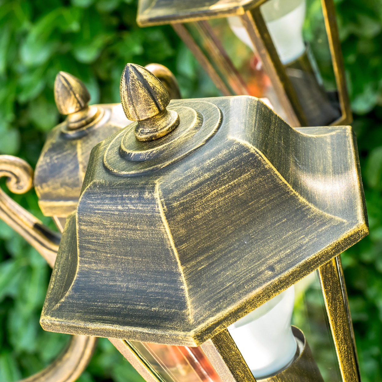 Nahaufnahme von verzierten, goldfarbenen Außenlampen. Die Lampen stehen vor einem Hintergrund aus grünem Laub, Details sichtbar.