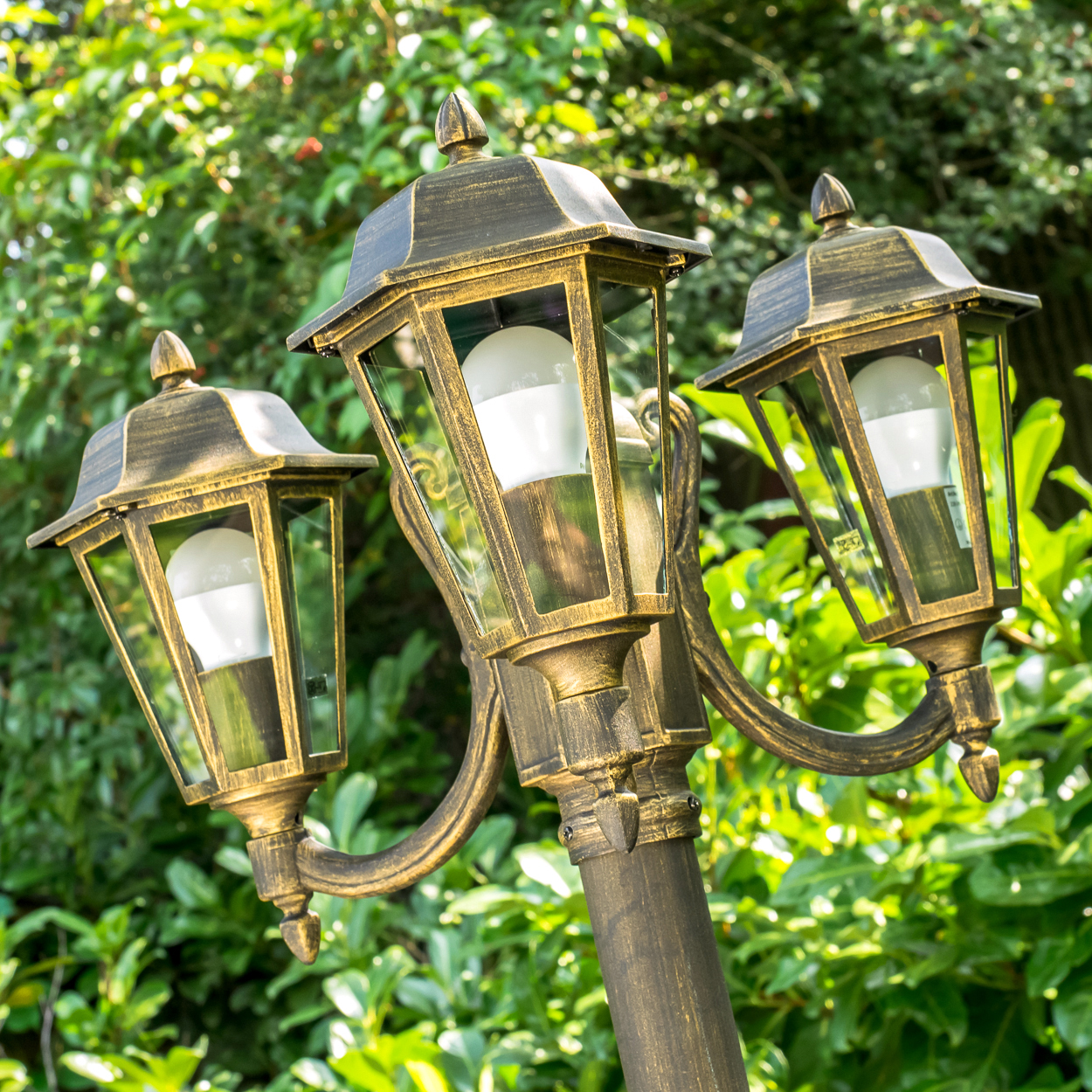 Eine bronzefarbene dreifache Laterne mit weißen LED-Lampen. Grünes Laub füllt den Hintergrund, wobei Sonnenlicht durch die Blätter scheint.